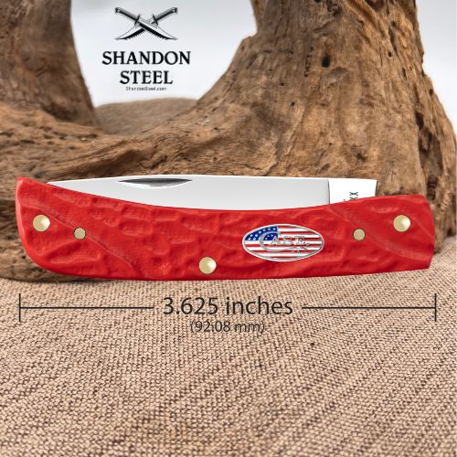 A red folding pocket knife with brass hardware and a silver blade, displayed on burlap with a wood background. The Shandon Steel logo is visible at the top, and a dimension overlay indicates the knife is 3.