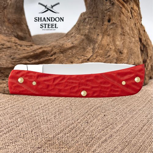 A red textured pocket knife with brass rivets and a white handle insert, displayed on a burlap surface with a piece of driftwood and a white background featuring the 'Shandon Steel' logo.