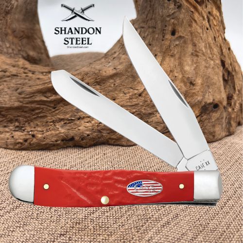 A red and silver folding pocket knife with two open blades, displayed on a burlap surface with a piece of driftwood and a branded card in the background. The brand 'Shandon Steel' is visible on the card.