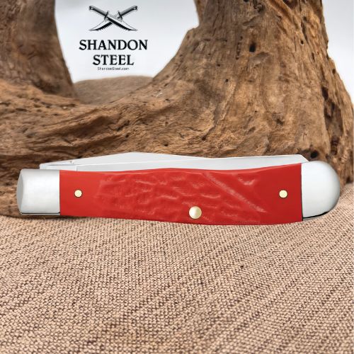 A red and silver folding pocket knife with brass pins rests on a textured burlap surface in front of a large piece of driftwood. The Shannon Steel logo is visible in the upper background.