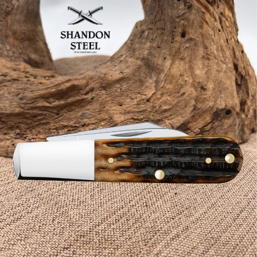 A folding pocket knife with a brown and black textured handle and a white bolster, displayed on a burlap surface with a piece of driftwood in the background. The brand logo 'SHANDON STEEL' is visible in the upper left.