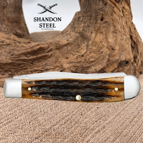 A folding pocket knife with a polished bone handle and white steel bolsters, displayed on a burlap surface with a piece of driftwood in the background. The brand logo 'SHANDON STEEL' is visible in the upper left.