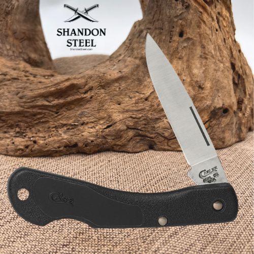 A folding pocket knife with a black textured handle and a silver steel blade, displayed on a burlap surface with a piece of driftwood in the background. The brand logo 'Shandon Steel' is visible in the upper left.