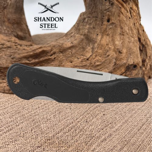 A folding pocket knife with a black textured handle and a silver metal blade, positioned horizontally in front of a rustic wood driftwood log and burlap fabric. The brand logo 'SHANDON STEEL' is visible in the upper left corner.