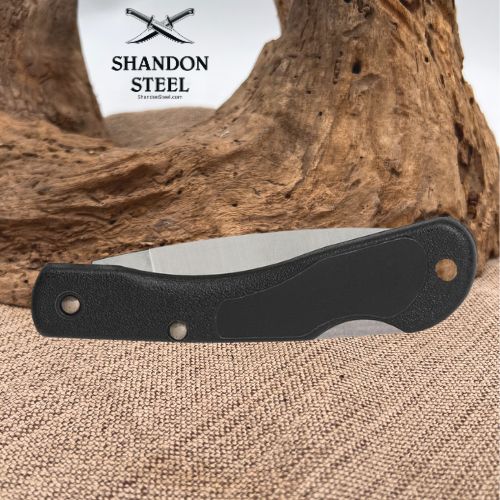 A folding pocket knife with a textured black handle and a silver steel blade, displayed horizontally on a burlap surface with a piece of driftwood in the background. The brand logo 'SHANDON STEEL' is visible in the upper left corner.