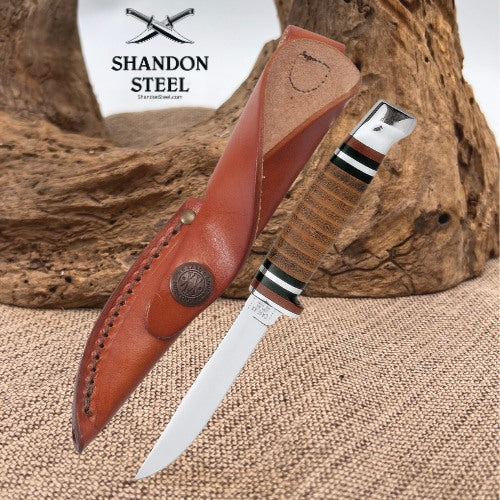 A fixed-blade knife with a silver blade and a brown wrapped handle, shown alongside a brown leather sheath. The background features a rustic wood log and burlap fabric, with the 'Shandon Steel' logo visible in the upper left.