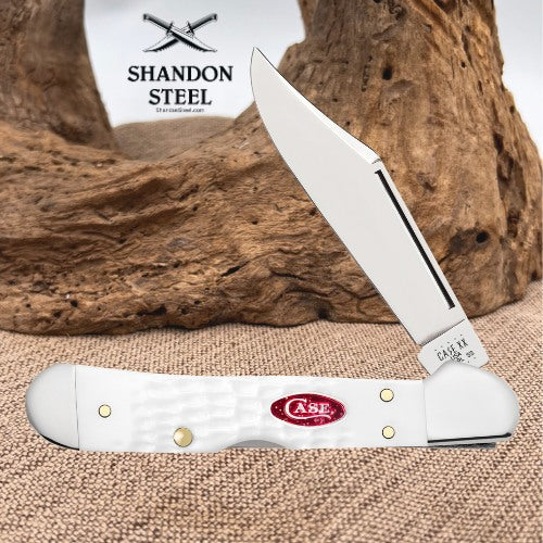 A white Case folding pocket knife with a silver blade, displayed open on a burlap surface with a piece of driftwood and a branded white card in the background. The knife features brass rivets and a red oval CASE logo.