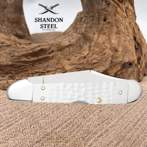 A white pocket knife with a textured handle and brass pins, displayed on a burlap surface with a piece of driftwood and a white background card showing the Shandon Steel logo.
