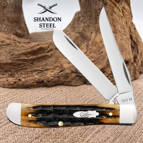 A double-blade pocket knife with polished silver blades and a carved handle made of brown and black bone with white end caps, displayed on a burlap surface with a piece of driftwood and a white Shandon Steel sign in the background.