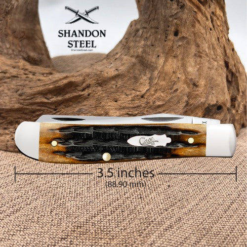 A folding pocket knife shown horizontally on a burlap surface with a large piece of driftwood behind it. The knife has a white handle with a black and brown wood inlay and brass pins.