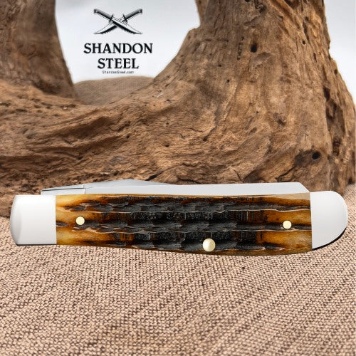 A folding pocket knife with a carved brown and black horn handle and white end caps, displayed on a burlap surface with a large piece of driftwood in the background. The Shandon Steel logo is visible in the upper left.