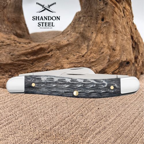 A folding pocket knife with a dark gray and black patterned handle and white end caps, displayed on a burlap surface with a piece of driftwood in the background. The brand logo 'SHANDON STEEL' is visible in the upper left.
