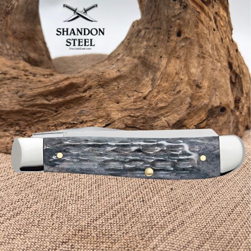 A folding pocket knife with a textured gray and black handle and silver-toned metal bolsters, displayed on a burlap surface with a large piece of driftwood in the background. The brand logo 'SHANDON STEEL' is visible in the upper left.