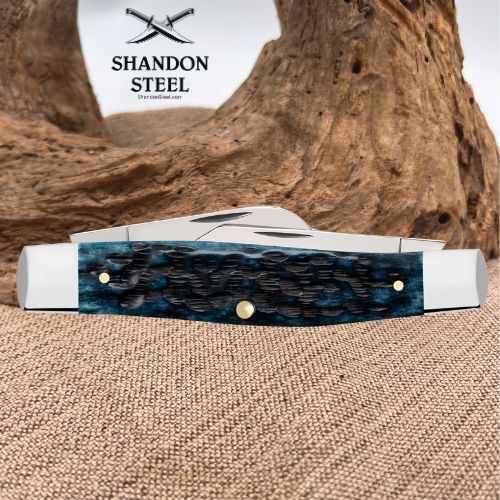 A folding pocket knife with a textured blue and black inlay handle and silver-toned metal blades, displayed on a burlap surface with a large piece of driftwood in the background. The brand logo 'SHANDON STEEL' is visible in the upper left.