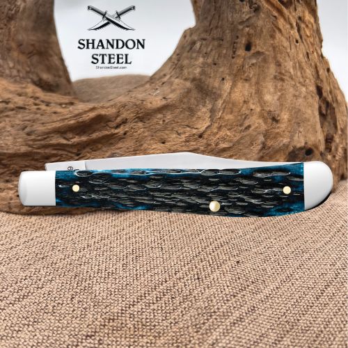 A folding pocket knife with a textured blue and black handle, resting on a burlap surface in front of a piece of driftwood. The brand logo 'SHANDON STEEL' is visible in the upper left corner.