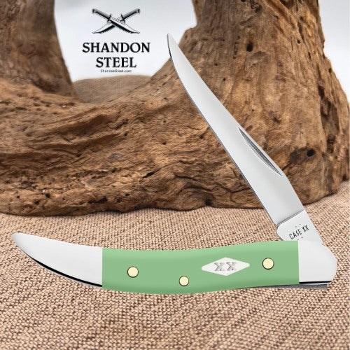 A folding pocket knife with a sage green handle and silver blades, displayed on a burlap surface with a piece of driftwood and a branded card in the background. The brand 'Shandon Steel' is visible on the card.