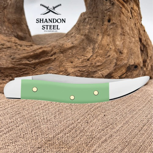 A folding pocket knife with a sage green handle and white metal bolster, displayed on a burlap surface with a piece of driftwood in the background. The Shandon Steel logo is visible in the upper left corner.