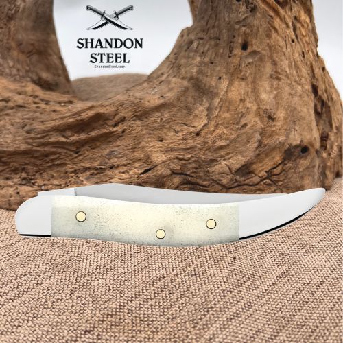 A folding pocket knife with a textured off-white handle and silver blade, resting on a burlap surface with a piece of driftwood in the background. The brand logo 'SHANDON STEEL' is visible in the upper left.
