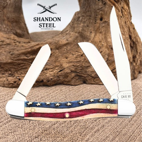 A folding pocket knife with two open blades, featuring a decorative handle with an American flag pattern of red, white, and blue, and gold stars. The knife is displayed on a burlap surface with a piece of driftwood in the background.