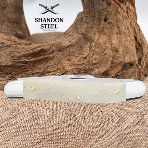 A folding pocket knife with a light gray handle and white end caps, displayed horizontally on a burlap surface with a large piece of driftwood in the background. The Shandon Steel logo is visible in the upper left corner.