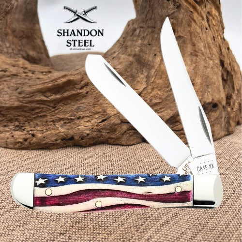 A folding pocket knife with a double-edged blade and a handle featuring an American flag pattern, displayed on a burlap surface with a piece of driftwood and a white Shandon Steel logo card in the background.
