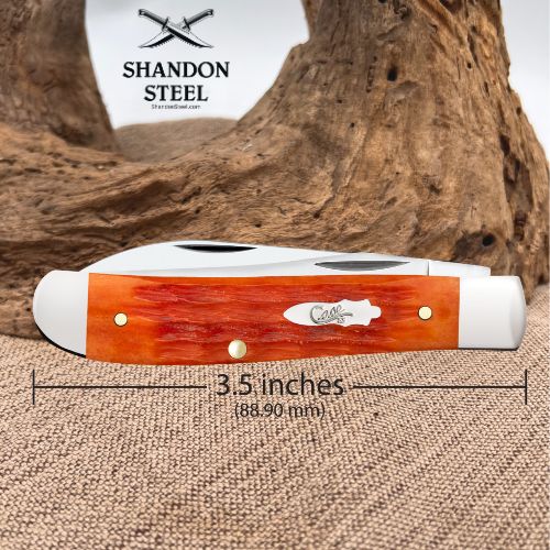 A folding pocket knife with a reddish-orange woodgrain handle and white metal blade components, shown on a burlap surface with a piece of driftwood behind it. A dimension line indicates the knife is 3.