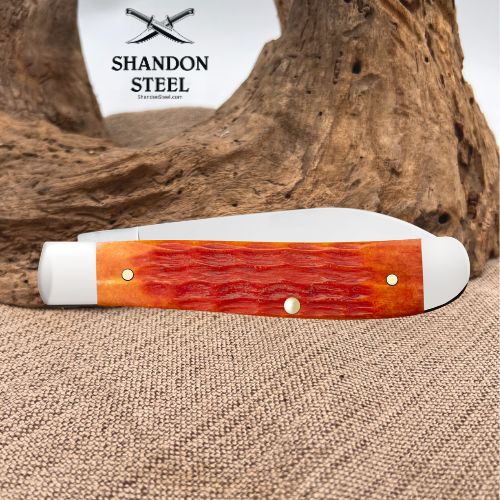 A folding pocket knife with a textured orange and red handle, displayed horizontally on a burlap surface with a piece of driftwood in the background. The brand logo 'SHANDON STEEL' is visible in the upper left corner.