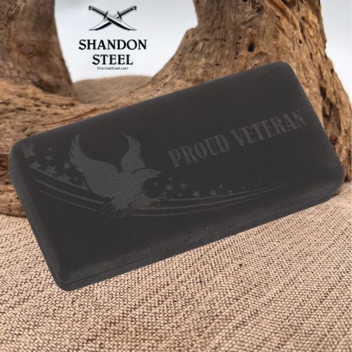 A black rectangular foam or rubber pad with a debossed eagle and stars design and the words 'PROUD VETERAN' on the front, shown on burlap with a tree root in the background. The brand name 'SHANDON STEEL' with crossed swords appears in the upper left.