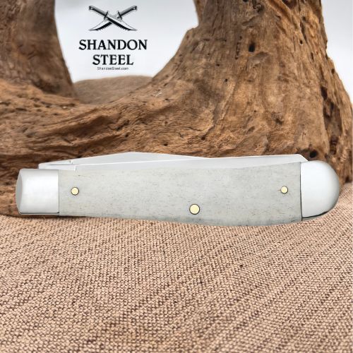 A folding pocket knife with a textured light gray handle and brass pins, resting on a burlap surface with a piece of driftwood in the background. The brand logo 'SHANDON STEEL' is visible in the upper background.