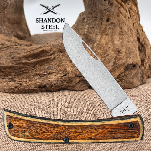 A folding pocket knife with a satin finish steel blade and a curved wooden handle, displayed on a burlap surface with a piece of driftwood in the background. The brand logo 'Shandon Steel' is visible in the upper left.