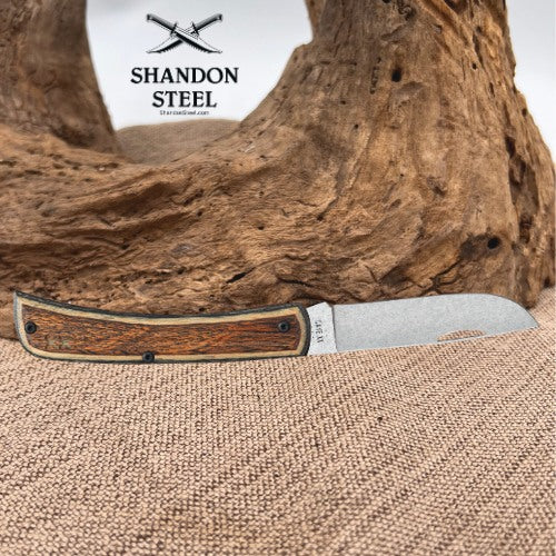 A folding pocket knife with a brown wood-grain handle and a silver steel blade, resting on a burlap surface in front of a piece of driftwood. The Shandon Steel logo is visible in the upper left corner.