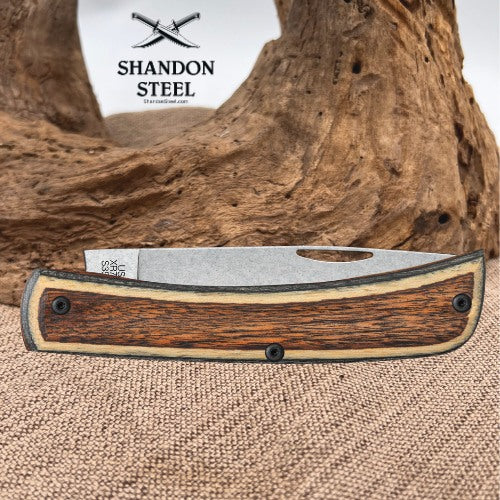 A folding pocket knife with a wood-grain handle and a silver steel blade, displayed on a burlap surface with a piece of driftwood in the background. The brand logo 'SHANDON STEEL' is visible in the upper left corner.