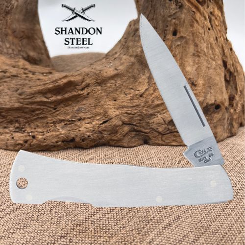 A folding pocket knife with a satin-finished silver blade and handle, displayed diagonally in front of a rustic piece of driftwood on burlap. The brand logo 'SHANDON STEEL' is visible in the upper left, and the brand name 'Shandon Steel' is engraved on the bolster of the knife.