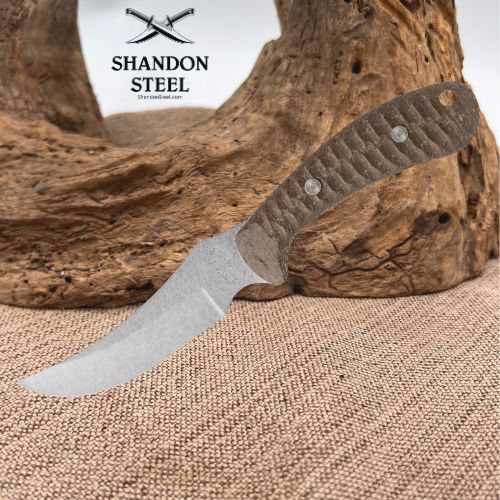 A fixed-blade knife with a textured gray handle and a silver steel blade, displayed diagonally on a rustic piece of driftwood over a burlap surface. The brand logo 'SHANDON STEEL' is visible in the upper left corner.