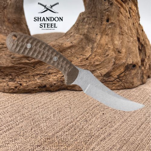 A fixed-blade knife with a textured brown handle and a satin-finished steel blade, resting on a piece of driftwood over a burlap surface. The brand logo 'SHANDON STEEL' is visible in the upper left.