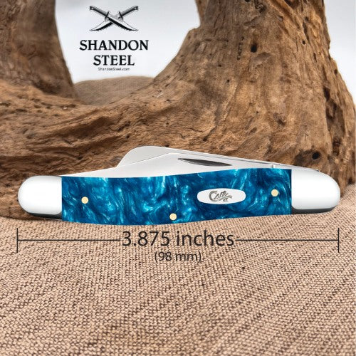 A folding pocket knife with a blue marbled resin handle and white end caps, shown on a burlap surface with a piece of driftwood behind it. A dimension line across the handle reads 3.
