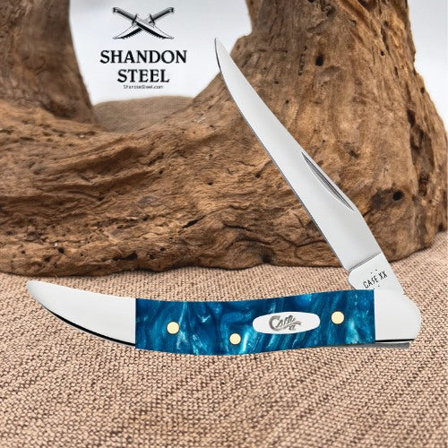 A folding pocket knife with a curved blue marbled handle and a silver blade, displayed on burlap in front of a piece of driftwood. The brand name 'SHANDON STEEL' is visible in the upper left.