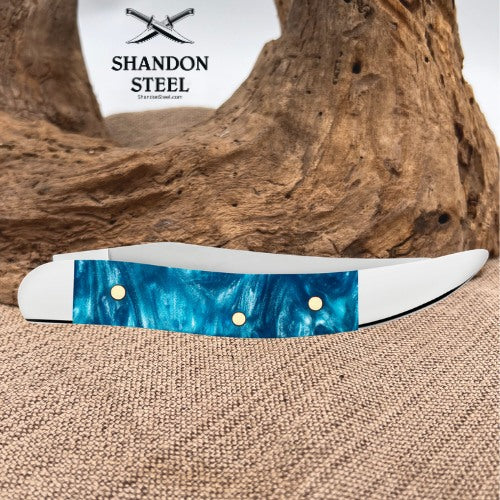 A folding pocket knife with a vibrant blue marbled inlay and white handle scales, displayed on a burlap surface with a piece of driftwood in the background. The brand logo 'SHANDON STEEL' is visible in the upper left.