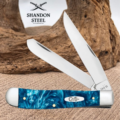 A folding pocket knife with a blue marbled handle and two silver blades, displayed on a burlap surface with a piece of driftwood and a white card reading SHANDON STEEL in the background.