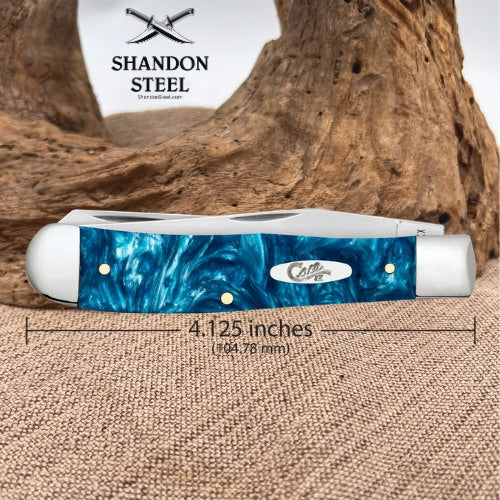 A folding pocket knife with a blue marbled handle and silver metal bolster, shown on a burlap surface with a piece of driftwood behind it. A dimension line indicates the knife is 4.