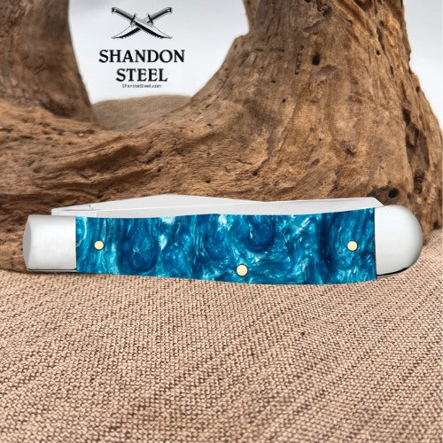 A pocket knife with a blue marbled resin handle and white bolster ends, displayed on a burlap surface with a piece of driftwood behind it. The brand logo 'SHANDON STEEL' is visible in the upper background.