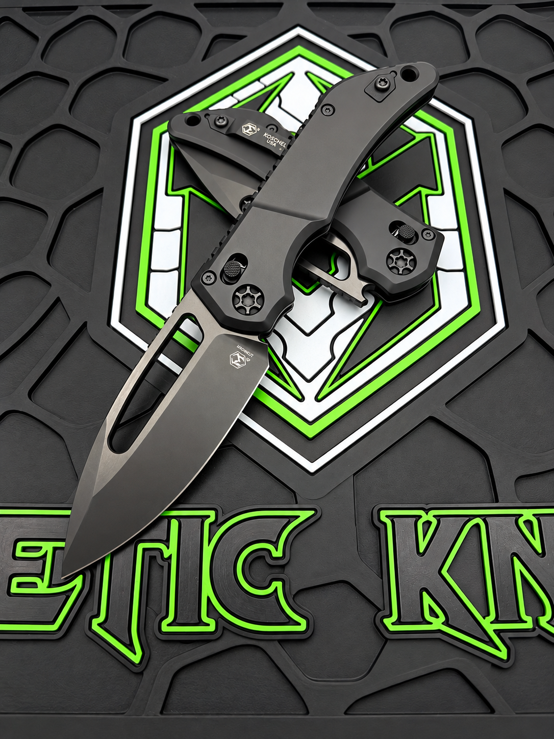 A black tactical folding knife with a matte finish is displayed open on a textured black mat featuring a green and white geometric logo and the partial brand name 'METIC KNA'.