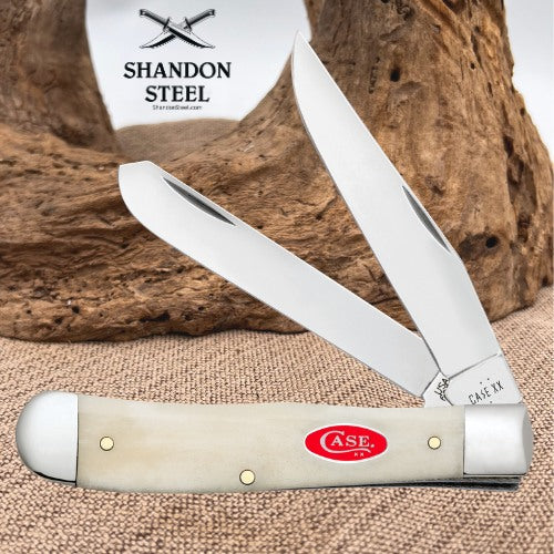 An open double-ended pocket knife with a cream-colored bone handle and polished steel blades, displayed on a burlap surface with a piece of driftwood and a branded card in the background. The handle features a red oval 'A.