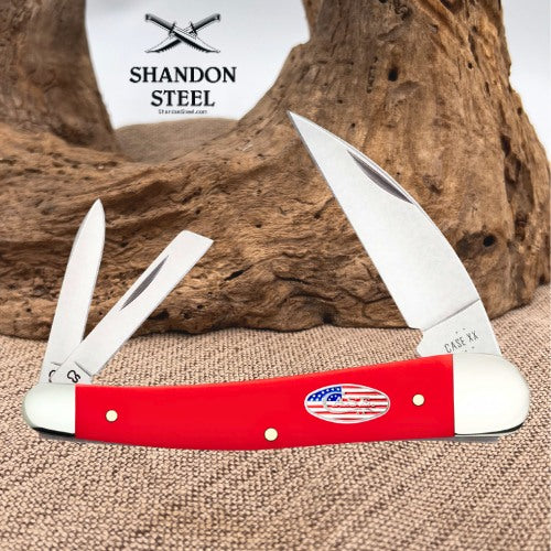 A red and white double-blade pocket knife with a small American flag emblem on the handle, displayed open on a burlap surface with a piece of driftwood and a white Shandon Steel sign in the background.