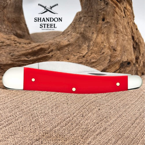 A red and white folding pocket knife with brass pins, displayed horizontally in front of a rustic wood driftwood log and burlap fabric background, with the Shandon Steel logo visible in the upper left.