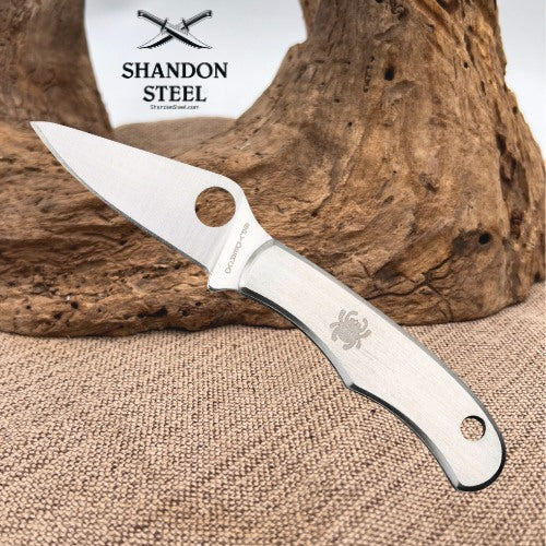 A stainless steel folding knife with a silver handle and blade, featuring a circular hole in the blade and an engraved spider logo on the handle. The knife is displayed on a textured burlap surface with a piece of driftwood in the background.