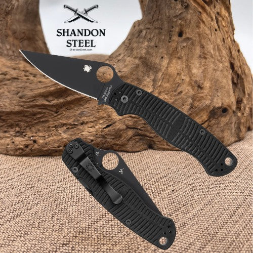 A black folding pocket knife with a textured handle and partially serrated blade, shown alongside its matching black sheath. The background features a rustic piece of driftwood and burlap fabric, with the 'Shandon Steel' logo printed at the top left.